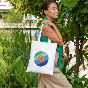 Puede incluir: Una bolsa de tela blanca con un diseño circular de colores que presenta una flor. El círculo tiene tonos de verde, azul y naranja.