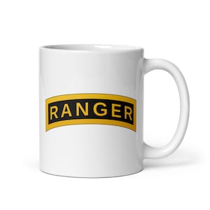 May include: A white ceramic mug featuring a curved rectangular patch design. The patch has a black background with a gold border and the word RANGER printed in bold gold capital letters centred on the front of the mug.