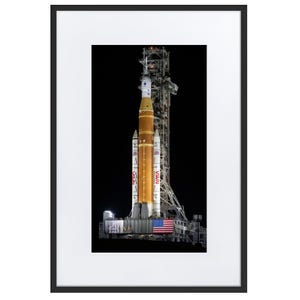 Op de afbeelding: Een ingelijste print met een NASA-raket op een lanceerplatform 's nachts. De raket is voornamelijk oranje en wit, met het NASA-logo zichtbaar. Een Amerikaanse vlag wordt getoond aan de basis van de lanceerstructuur.