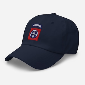 Può includere: Un cappellino da baseball blu navy con una toppa ricamata sul davanti. La toppa presenta un quadrato rosso con bordo blu, contenente le lettere bianche AA e un contorno circolare bianco, sormontato da una linguetta blu con la parola AIRBORNE.