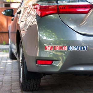 以下が含まれることがあります： 「NEW DRIVER BE PATIENT」と書かれた赤と白のステッカーが貼られた灰色の車のリアバンパー。車のテールライトは赤色で、バンパーには赤いリフレクターが付いています。車はレンガの表面に駐車されています。