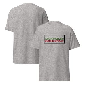 Puede incluir: Una camiseta gris con un rectángulo negro que contiene el texto "TASK FAILED SUCCESSFULLY" en letras verdes y rojas.