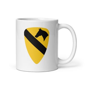 Puede incluir: Una taza de cerámica blanca con un emblema centrado en forma de escudo. El emblema tiene un fondo amarillo con una franja negra diagonal y una silueta negra de una cabeza de caballo situada en la parte superior derecha del diseño.