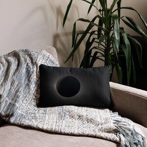 May include: A black rectangular pillow with a large, dark circle design. The circle has a subtle, glowing outline. The pillow is placed on a patterned blanket and a neutral-toned sofa, creating a modern aesthetic.
