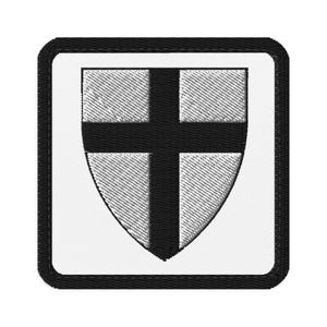 May include: Embroidered patch with a shield design. It features a black cross on a white background, enclosed within a black-outlined shield and a square frame. The patch has a textured appearance.