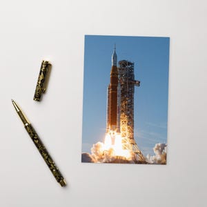 May include: A photograph of a rocket launching into the sky, with flames and smoke billowing from the base. A black and gold pen and pen cap with a floral design are to the left of the photo. The pen is approximately 5.5 inches long.