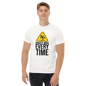 Pode incluir: Camiseta branca com um gráfico de sinal de advertência amarelo. O sinal tem uma figura de pau caindo e o texto "GRAVITY WINS EVERY TIME".