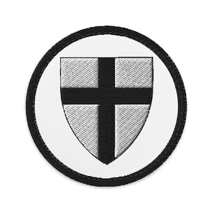 May include: Embroidered patch featuring a shield with a black cross on a white background, encircled by a black border. The shield has a textured, diagonal pattern. The patch is circular in shape.