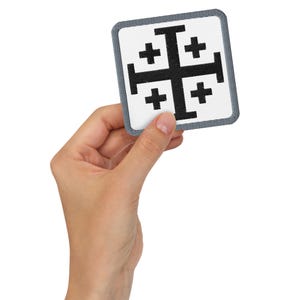 May include: A square patch featuring a black Jerusalem cross on a white background, with a grey border. The patch is held in a hand.