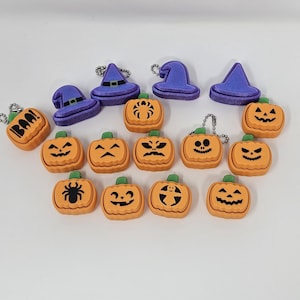 Könnte beinhalten: Eine Sammlung von Halloween-Themen-Anhängern und Schlüsselanhängern. Das Bild zeigt orangefarbene Kürbis-Anhänger mit geschnitzten Gesichtern und grünen Stielen, sowie lila Hexenhut-Anhänger. Einige Anhänger tragen das Wort "BOO!".