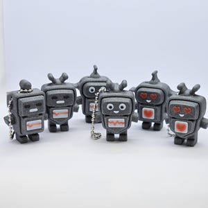 May include: Six small gray robot keychains with different facial expressions. Some robots have hearts on their chests and hearts for eyes.