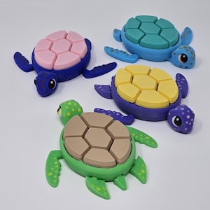 Könnte beinhalten: Vier bunte Spielzeug-Meeresschildkröten mit segmentierten Schalen. Jede Schildkröte hat eine andersfarbige Schale: rosa, blau, gelb und braun. Die Schildkröten sind aus Kunststoff und haben bemalte Details.