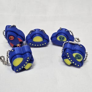 May include: Five blue keychains with different space-themed designs. The keychains feature a rocket, a UFO, and three alien faces. The keychains are made of a soft, pliable material.