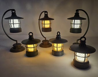 May include: Five decorative metal lanterns with a warm, rustic finish. Each lantern has a clear glass panel and a warm white LED light. The lanterns are arranged on a white surface.