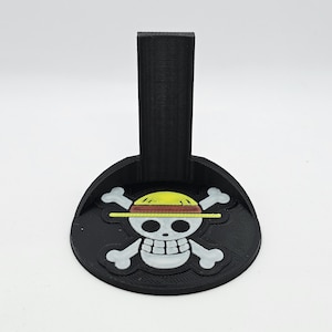 May include: A black 3D printed stand with a skull and crossbones design. The skull is white with a yellow hat and a red band. The stand has a vertical post for displaying a figure.