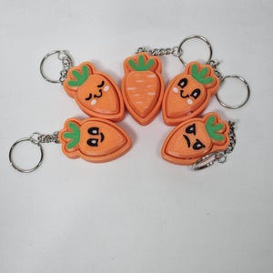 May include: Five orange carrot keychain charms with different facial expressions. Each charm has a green leafy top and a silver key ring.