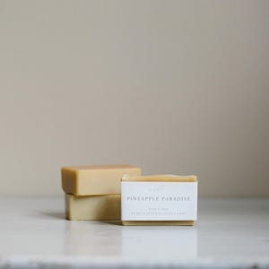 May include: Three rectangular bars of soap, two stacked and one in front with a label. The label reads "Pineapple Paradise" with additional text. The soap is a light yellow color, and the background is a neutral beige.