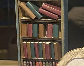 May include: Miniature bookshelf with a gold-colored ornate top and blue sides. The shelves are filled with tiny books in various colors, including red, brown, and green. A rolled-up scroll and other small items are on top.