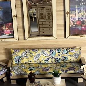 Könnte beinhalten: Miniatur-Wohnzimmerszene mit einem gelb geblümten Sofa, einem kleinen Couchtisch und gerahmten Kunstwerken. Ein Spiegel spiegelt einen Schrank wider. Eine kleine Pflanze und Bücher stehen auf dem Tisch, vor hellem Holzgrund.