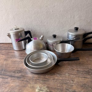 May include: A collection of vintage aluminum cookware including a percolator, a tea kettle, a lidded pot, a small bowl, and a set of nested round pans. Each piece features a metallic silver finish and black plastic handles or knobs.