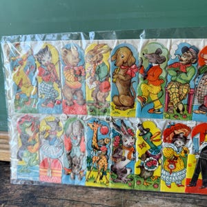May include: A collection of vintage paper die-cut animal characters in a plastic sleeve. The colorful illustrations include a cat playing a violin, a bear holding flowers, a rabbit with a letter, and a dog playing a flute. The characters are dressed in various outfits.