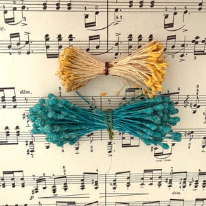 May include: Two bundles of craft supplies: one with yellow flower heads and natural stems, the other with turquoise flower heads. Both are tied with twine. The background is sheet music.