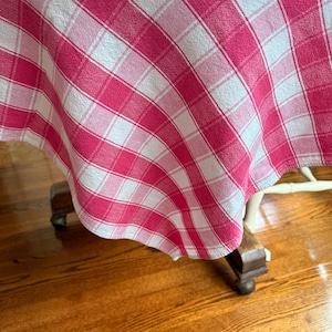 May include: A fabric tablecloth featuring a diagonal plaid pattern in shades of bright pink, light pink, and white. The textile has a visible woven texture and drapes over the edges of a surface, displaying a consistent grid of intersecting lines.