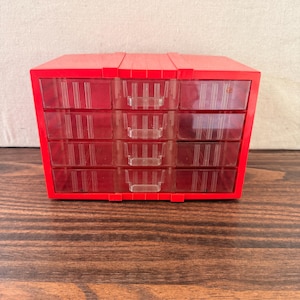 May include: A bright red plastic storage organizer featuring twelve individual clear drawers arranged in a four-by-three grid. Each drawer has a small integrated handle and vertical ribbed detailing on the front face of the transparent plastic.