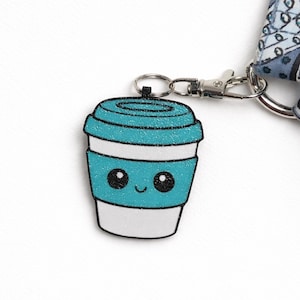 May include: A teal glitter coffee cup charm with a smiling face, black eyes, and a white base. The charm is attached to a silver-colored clasp and ring. The background is white.