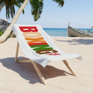 May include: A white beach towel draped over a wooden lounge chair. The towel features the word BOXING printed in large, stacked, multi-colored letters including red, orange, green, and brown, with the text USA Est. 2025 in red below the word.
