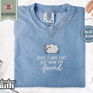 May include: A blue jean coloured t-shirt featuring an embroidered white sheep graphic and the text "ONCE I WAS LOST BUT NOW I'M found" in white thread. The shirt has a crew neckline and a Comfort Colors brand label on the inside collar area.