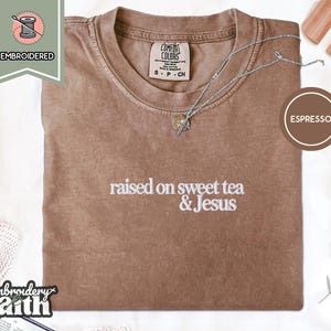 May include: An espresso-coloured short-sleeve t-shirt with white embroidered text that reads raised on sweet tea & Jesus. The shirt features a crew neckline and a Comfort Colors brand label on the inside collar, with a silver chain necklace resting on top.