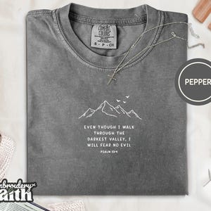 May include: A charcoal grey short-sleeve t-shirt featuring a white line art graphic of mountain peaks and birds. Below the mountains, the text reads: EVEN THOUGH I WALK THROUGH THE DARKEST VALLEY, I WILL FEAR NO EVIL PSALM 23:4. A silver chain necklace rests on the collar.