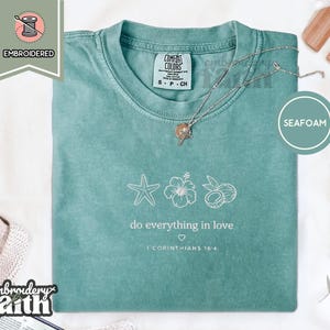 Puede incluir: Una camiseta de manga corta color verde espuma de mar con bordado blanco de una estrella de mar, una flor de hibisco y un coco. Debajo de los iconos, el texto dice "do everything in love" con un pequeño corazón y "1 CORINTHIANS 16:14".