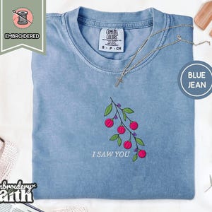 May include: A blue jean colored t-shirt with a crew neckline and a Comfort Colors brand label. The shirt features an embroidered design of a green leafy branch with pink berries and the white text I SAW YOU positioned below the branch.