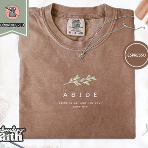 May include: An espresso-colored t-shirt featuring an embroidered green leafy branch graphic above the white text ABIDE and the verse ABIDE IN ME, AND I IN YOU. JOHN 15:4. The shirt has a crew neckline and a Comfort Colors brand label inside.