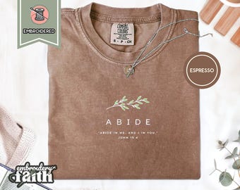 May include: An espresso-coloured t-shirt featuring an embroidered green leafy branch graphic above the white text ABIDE and the verse ABIDE IN ME, AND I IN YOU. JOHN 15:4. The shirt has a crew neckline and a Comfort Colors brand label inside.