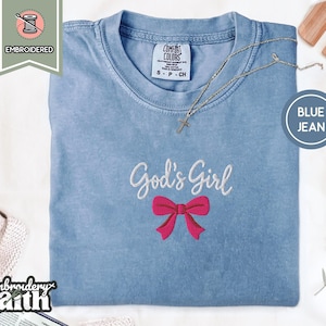 Puede incluir: Una camiseta de manga corta color azul jean con texto bordado en blanco que dice God's Girl sobre un lazo bordado de color rosa brillante. La camiseta tiene un cuello redondo y una etiqueta de la marca Comfort Colors visible en el cuello.
