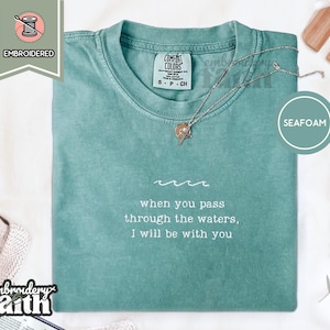 Könnte beinhalten: Ein kurzärmeliges T-Shirt in Meerschaumgrün mit weißem Sticktext, der "when you pass through the waters, I will be with you" lautet, unter einer kleinen gestickten Wellengrafik. Das T-Shirt hat einen Rundhalsausschnitt und ein Etikett.