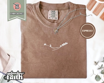 May include: An espresso-coloured short-sleeve t-shirt with a crew neckline. The shirt features a small white embroidered graphic on the centre chest depicting a shepherd with a staff and a flock of sheep on a curved line. A silver chain necklace rests on the collar.