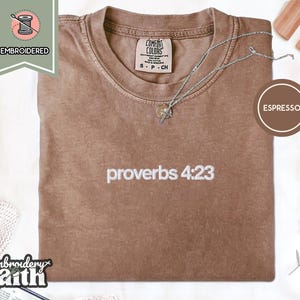 Könnte beinhalten: Ein espressofarbenes Kurzarm-T-Shirt mit weißem, gesticktem Text proverbs 4:23 mittig auf der Brust. Das T-Shirt hat einen Rundhalsausschnitt und ein Etikett der Marke Comfort Colors im Kragen, das die Größe S angibt.