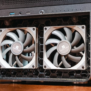 May include: A computer case featuring two cooling fans. The fans are grey and white, with multiple blades and a central logo. The case is black and has a honeycomb pattern.
