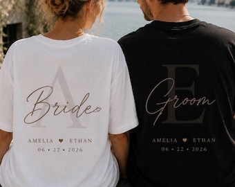 May include: A white t-shirt with the text Bride, a large letter A, the names Amelia and Ethan, and the date 06 22 2026, alongside a black t-shirt with the text Groom, a large letter E, the names Amelia and Ethan, and the date 06 22 2026.