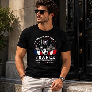 May include: A black short-sleeved t-shirt featuring a graphic print with the text World Cup 2026, France, and Pride Passion Football. The design includes the Eiffel Tower, the French Football Federation logo, a soccer ball, and the French flag.