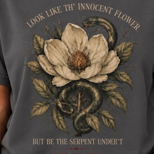 Puede incluir: Una camiseta gris carbón con un estampado gráfico de una gran flor blanca con una serpiente oscura enroscada alrededor de su tallo y hojas. El texto dice: LOOK LIKE TH' INNOCENT FLOWER, BUT BE THE SERPENT UNDER'T, MACBETH ACT 1, SCENE V.