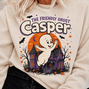 May include: A beige crewneck sweatshirt featuring a graphic print of Casper the Friendly Ghost in front of a purple haunted house and a large orange moon. The design includes black bats, orange and purple stars, and the text The Friendly Ghost Casper.