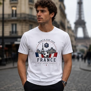 May include: A white short-sleeved t-shirt featuring a graphic print that includes the text World Cup 2026, France, and Pride Passion Football. The design displays a soccer ball, the Eiffel Tower, and a French flag motif in red, white, and blue.