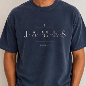 Könnte beinhalten: Ein marineblaues T-Shirt mit kurzen Ärmeln und einem weißen Grafikdruck. Das Design zeigt ein kleines Kreuz über dem Wort JAMES, gefolgt von dem Text A SERVANT OF GOD und der Referenz JAMES 1:1, alles zentriert in einer klaren Serifenschrift.