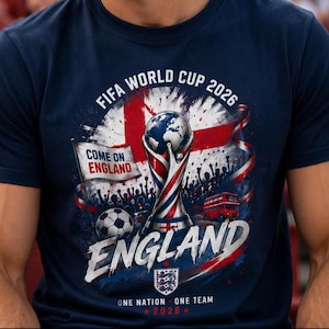 Könnte beinhalten: Ein marineblaues T-Shirt mit einer Grafik des FIFA World Cup 2026 Pokals, einem Fußball, einem roten Doppeldeckerbus und der englischen Flagge. Der Text lautet FIFA WORLD CUP 2026, COME ON ENGLAND, ENGLAND sowie ONE NATION ONE TEAM 2026.