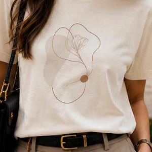 May include: A cream-colored short-sleeve t-shirt featuring a brown line art graphic of a flower and abstract shapes on the front. The design includes a thin, continuous line drawing of a floral stem and a solid brown circle accenting the composition.